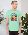 Man wearing a light mint Milk Sandwich t-shirt featuring a colorful illustration of six diverse cartoon kids smiling under a rainbow with the text "LET'S BE FRIENDS" standing indoors next to large green plants.