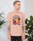 Man wearing a light peachy Milk Sandwich t-shirt featuring a colorful illustration of six diverse cartoon kids smiling under a rainbow with the text "LET'S BE FRIENDS" standing indoors next to large green plants.
