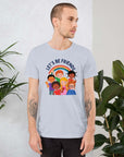 Man wearing a light blue Milk Sandwich t-shirt featuring a colorful illustration of six diverse cartoon kids smiling under a rainbow with the text "LET'S BE FRIENDS" standing indoors next to large green plants.
