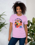 Smiling woman wearing a lilac Milk Sandwich t-shirt with a colorful "LET'S BE FRIENDS" graphic featuring six diverse cartoon kids and a rainbow, standing indoors near leafy green plants.