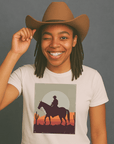 Smiling person with dreadlocks wearing a cowboy hat and winking, dressed in a light-colored t-shirt featuring a graphic of a cowboy riding a horse at sunset with cacti in the background.