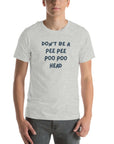 Man wearing light grey “DON’T BE A PEE PEE POO POO HEAD” t-shirt, standing casually with hands by pockets.