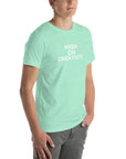 Young man wearing a mint t-shirt with white text ‘HIGH ON CREATIVITY’, standing angled to the side with hands in pockets.