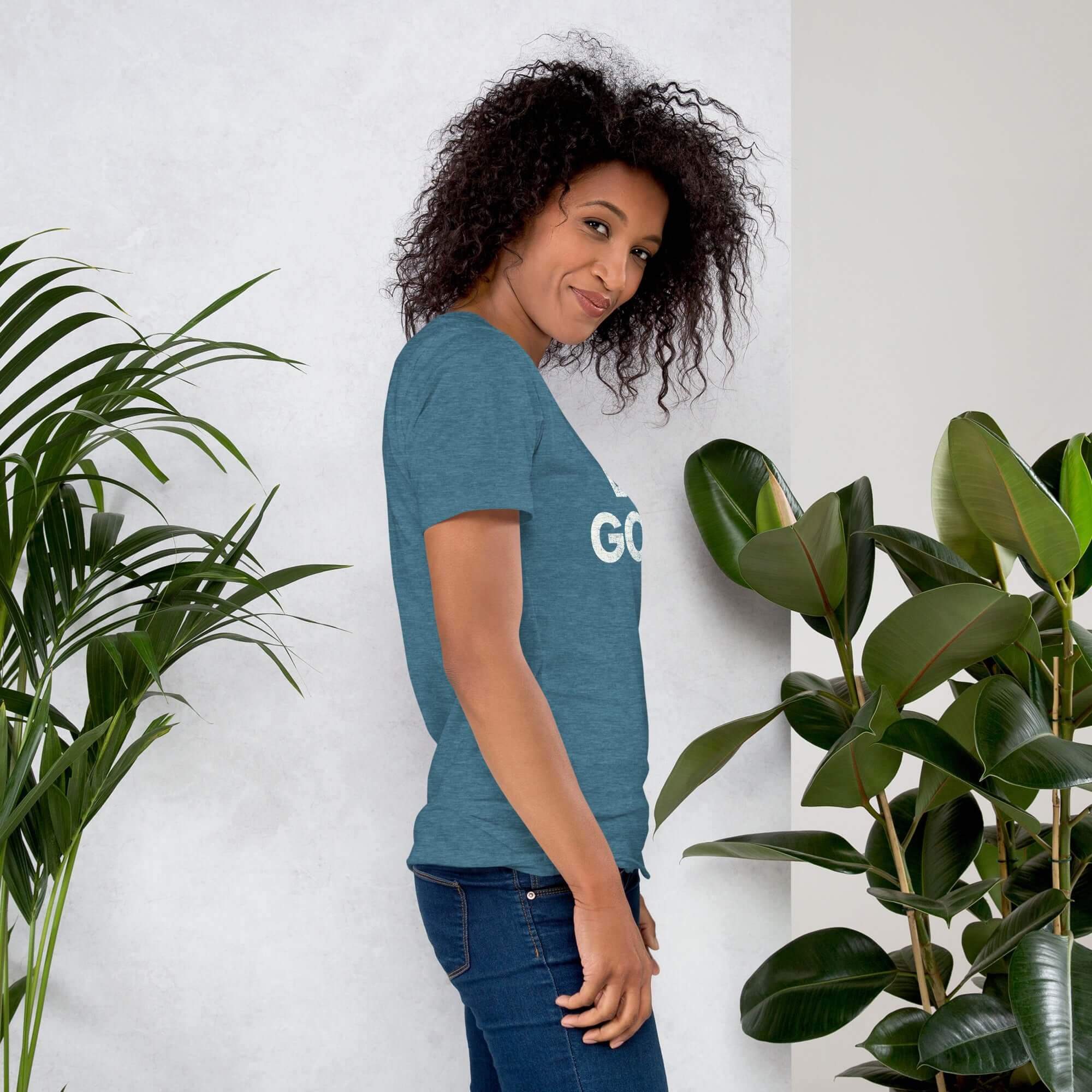 Side profile of smiling woman in teal Milk Sandwich T-shirt with DO GOOD text, standing near plants, casual inspirational graphic tee.