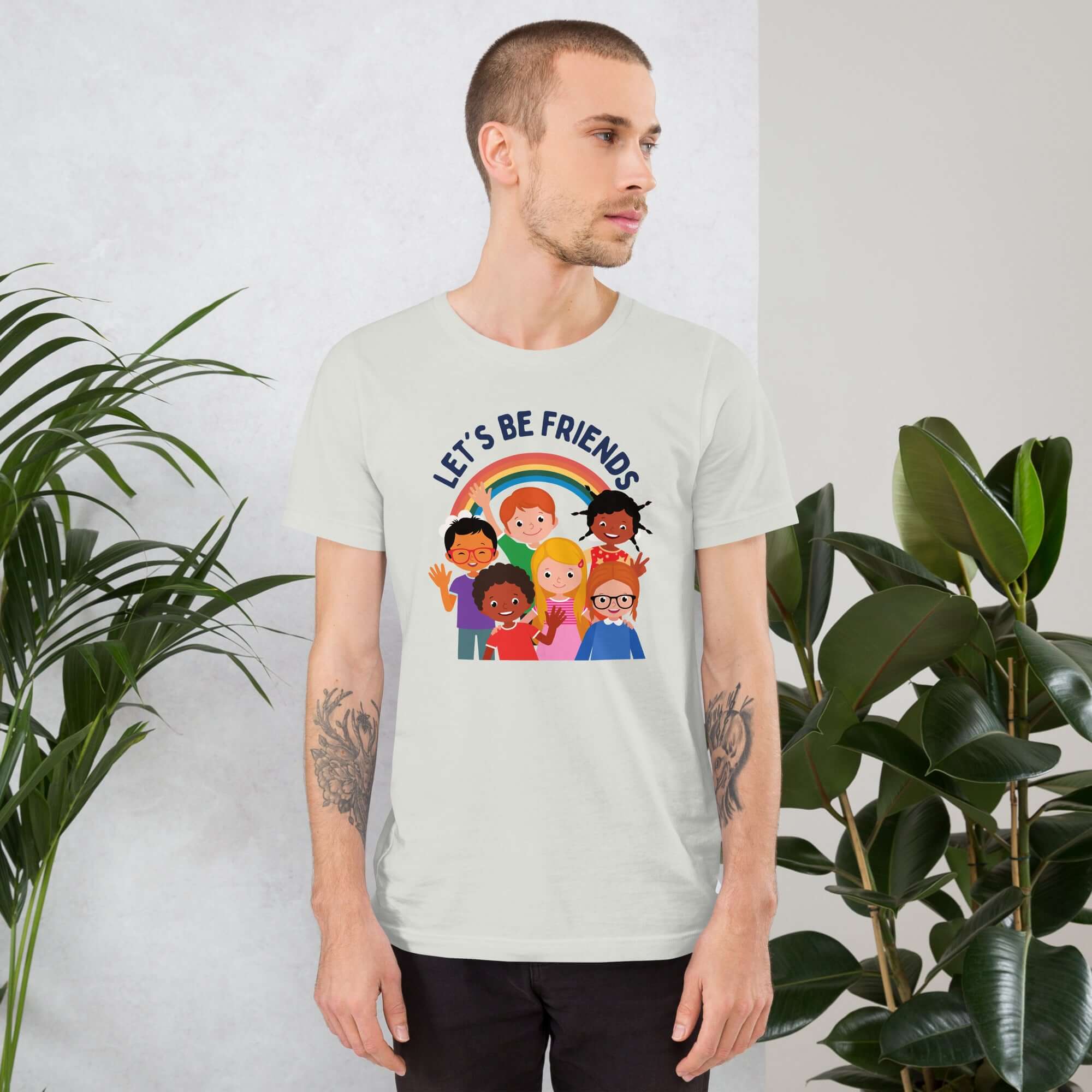 Man wearing a ash Milk Sandwich t-shirt featuring a colorful illustration of six diverse cartoon kids smiling under a rainbow with the text &quot;LET&#39;S BE FRIENDS&quot; standing indoors next to large green plants.