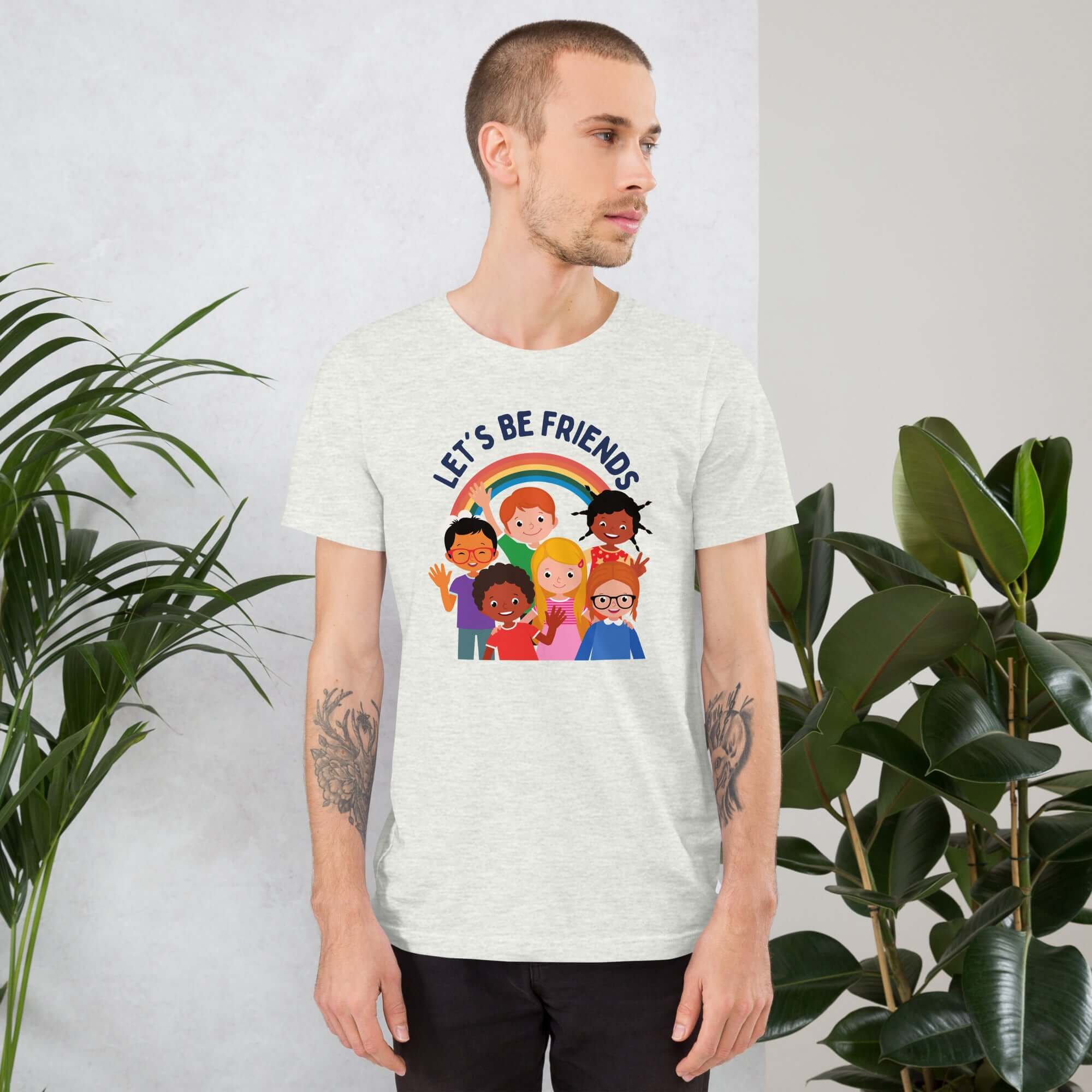 Man wearing a silver Milk Sandwich t-shirt featuring a colorful illustration of six diverse cartoon kids smiling under a rainbow with the text &quot;LET&#39;S BE FRIENDS&quot; standing indoors next to large green plants.
