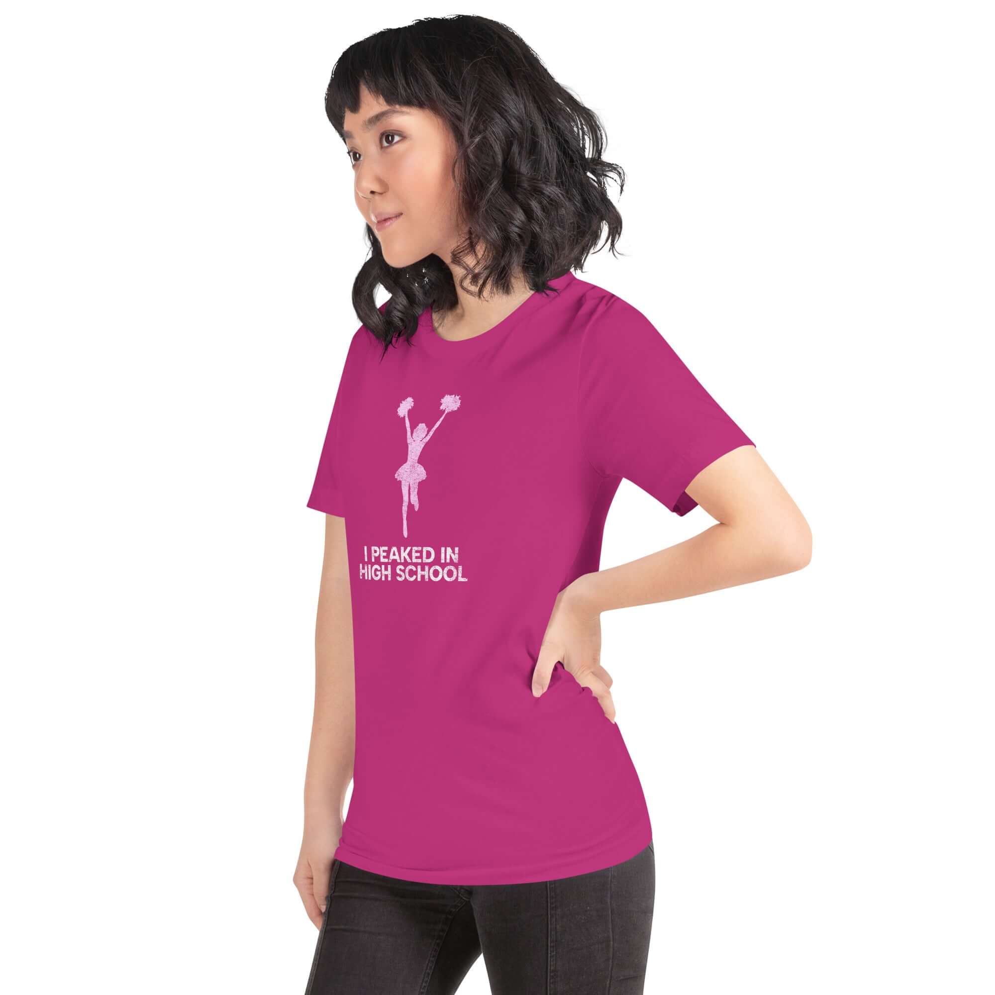 Model wearing a vibrant pink unisex t-shirt that says &quot;I Peaked in High School&quot; with a cheerful cheerleader graphic.