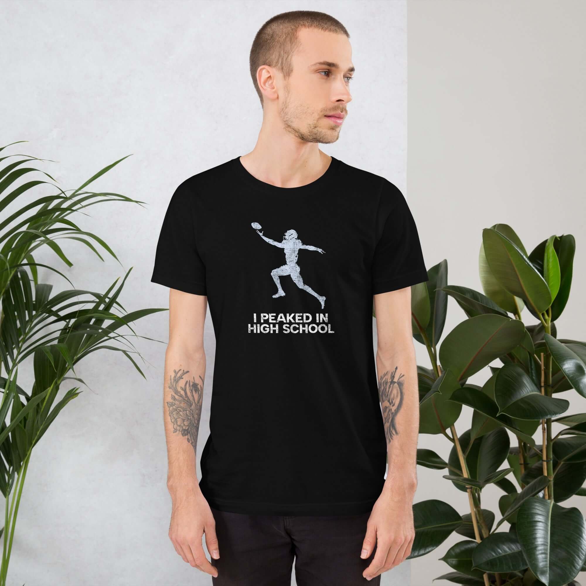 Unisex &quot;I Peaked In High School Football&quot; t-shirt worn by a model, surrounded by greenery, perfect for nostalgic sports fans.